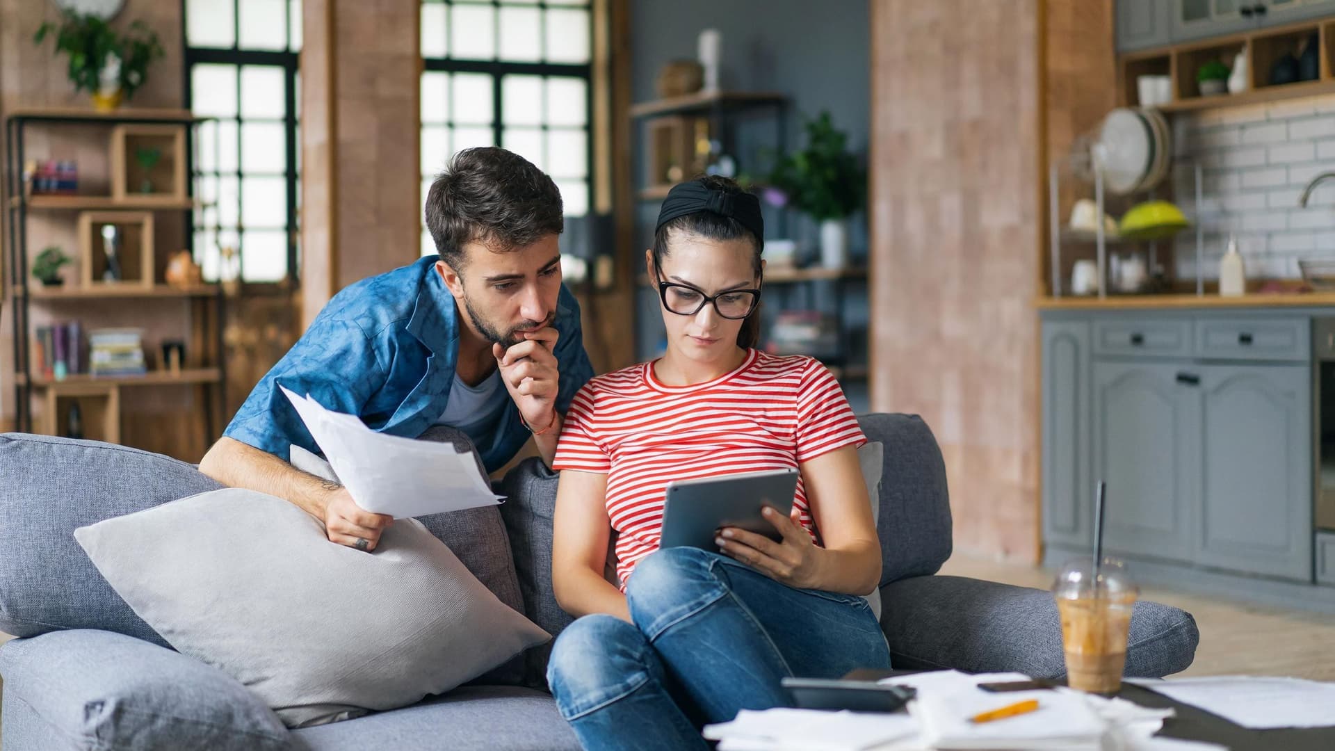 Ein Paar sitzt zusammen auf einem Sofa in einem gemütlichen Wohnraum. Die Frau trägt eine große Brille und schaut konzentriert auf ein Tablet, während der Mann neben ihr steht, Unterlagen in der Hand hält und nachdenkt.